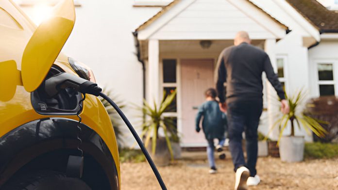 Cette image montre une voiture électrique jaune en train de se recharger à l'extérieur d'une maison, tandis qu'un homme et un enfant se dirigent vers la porte d'entrée. La scène évoque une vie familiale moderne et durable, mettant l'accent sur l'adoption de technologies écologiques telles que les véhicules électriques.
